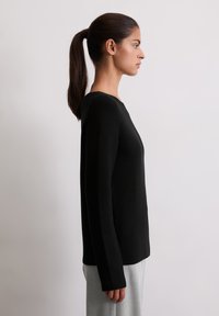 Tânără cu p�ărul închis la culoare prins în coadă de cal, îmbrăcată cu un top lung negru și pantaloni gri deschis, stând de profil pe un fundal simplu.