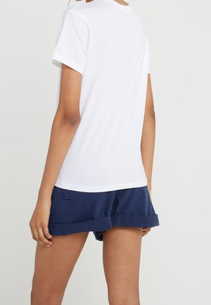 Persoon die een effen wit T-shirt met korte mouwen en marineblauwe opgerolde korte broek draagt, gezien van achter tegen een lichte achtergrond.