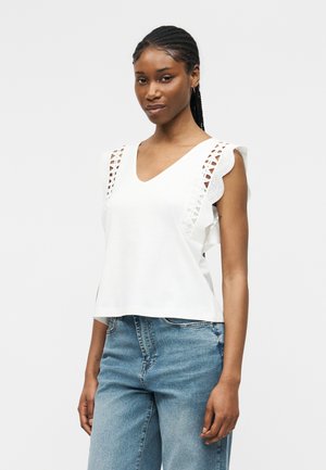 Ženska s kitkami v laseh, oblečena v belo brezrokavnik z čipkastimi detajli, kombiniranega s svetlo modrimi visokim pasom kavbojkami na enobarvnem ozadju.