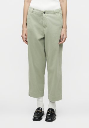 Άτομο που φοράει ανοιχτοπράσινο παντελόνι με φαρδιά παντελόνια μέχρι τον αστράγαλο, λευκές κάλτσες, μαύρα loafers και λευκό ριμπ μπλουζάκι, στέκεται μπροστά σε απλό φόντο.