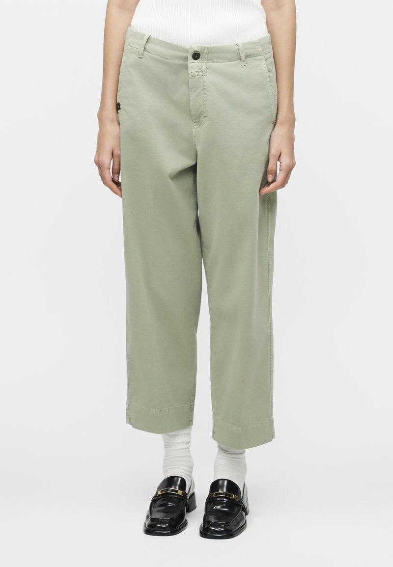 Άτομο που φοράει ανοιχτοπράσινο παντελόνι με φαρδιά παντελόνια μέχρι τον αστράγαλο, λευκές κάλτσες, μαύρα loafers και λευκό ριμπ μπλουζάκι, στέκεται μπροστά σε απλό φόντο.