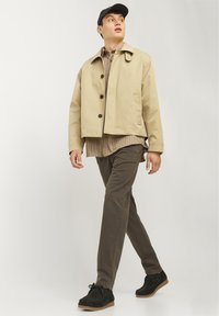 Beige short jacket with black buttons, worn over a plaid shirt. Dark green pants and black shoes. Simple design and casual fit.