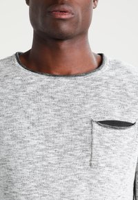 Maglione grigio mélange con scollatura rotonda, lavorazione a maglia testurizzata e una piccola tasca nera sul lato sinistro del petto. Cuciture nere a contrasto visibili.