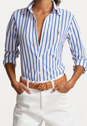 Camisa blanca de botones con rayas verticales azules, mangas largas arremangadas en los puños. Combinada con un cinturón trenzado marrón y pantalones blancos.