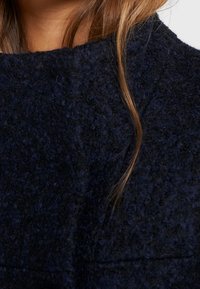 Tessuto blu navy con una trama texture e un aspetto peloso, messo in risalto da un ampio colletto e dettagli di cucitura visibili. Ciocche di capelli incorniciano parzialmente il colletto.