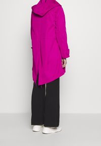 Fuchsia lange jas met een capuchon, voorzien van een getailleerde taille en splitten aan de zijkanten. Draagt over zwarte wijde broekspijpen en witte sneakers.