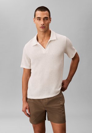 Mężczyzna ubrany w białą teksturowaną koszulkę polo z krótkim rękawem i brązowe szorty Calvin Klein na jednolitym szarym tle.
