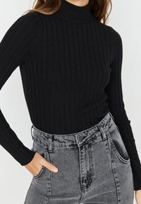 Schwarzer, gerippter Rollkragenpullover mit langen Ärmeln, kombiniert mit hochgeschnittenen grauen Jeansshorts, die über Knopf- und Taschendetails verfügen.