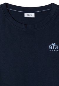 Camiseta azul marino oscuro con cuello redondo y un pequeño logo blanco bordado "BE KIND" con una palmera en la parte superior izquierda.