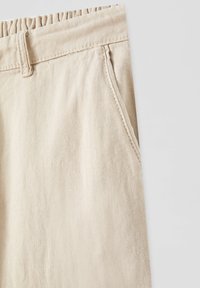 Jasne beżowe spodnie z elastycznym paskiem w talii, bocznymi kieszeniami i gładkim, teksturowanym materiałem. Zawierają proste detale stębnowania.