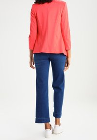 Persoon die een koraalroze blazer, blauwe jeans en witte sneakers draagt, staat met de rug naar de kijker op een witte achtergrond.