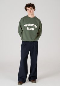 Jaunas vyras su garbanotais plaukais, dėvintis alyvuogių žalios spalvos džemperį su užrašu "PROHIBITED BERLIN", jūrinio stiliaus plačiomis kelnėmis ir rudos spalvos batais.