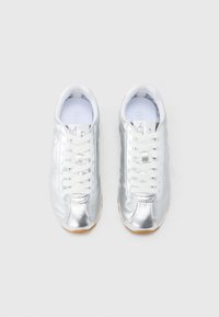 Chaussures de sport argentées avec une tige métallique brillante, des lacets blancs et une semelle en caoutchouc texturée. Discrète marque sur la languette.