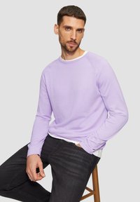 Chemise à manches longues de couleur violet clair avec un col rond et une texture côtelée, associée à un jean foncé. Modèle assis sur un tabouret en bois.