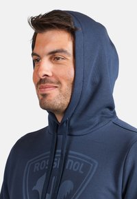 Felpa navy realizzata in morbido tessuto con una grafica logo frontale. Presenta un cappuccio ampio con cordino e una texture liscia, con vestibilità regolare.