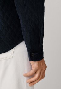 Ciemnoniebieska koszula z korduroy w diamentowy wzór, z mankietem na guziki i gładką fakturą, zestawiona z jasnymi spodniami.