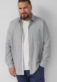 Camicia a maniche lunghe a quadri grigia con taschino frontale, colletto alla coreana e una maglietta bianca sotto. Tessuto liscio.