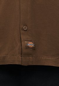 Τμήμα καφέ πουκάμισου Dickies που δείχνει κουμπί και ραμμένη ετικέτα της μάρκας στο τελείωμα.