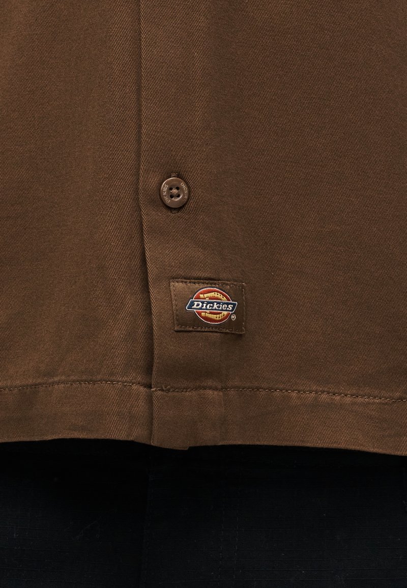 Sección de camisa Brown Dickies que muestra el botón y la etiqueta de la marca cosida en el dobladillo.