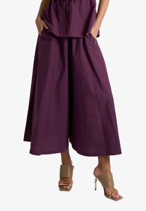 Donna con pantaloni viola a gamba larga con tasche e sandali beige con tacco alto e dettagli dorati, in piedi con le mani nelle tasche.