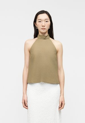 Młoda kobieta o długich ciemnych włosach, ubrana w oliwkowy top na ramiączkach z ozdobnym wiązaniem i teksturowaną białą spódnicę, stoi na tle jednolitego jasnego tła.