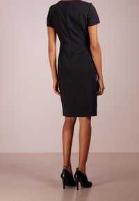 Femme se tenant de dos, portant une robe noire ajustée arrivant au genou avec des manches courtes et des chaussures à talons hauts noires.