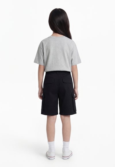 Enfant aux longs cheveux foncés, debout de dos, portant un t-shirt gris à manches courtes, un short cargo noir, des chaussettes blanches et des baskets blanches.