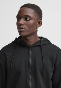 Black zip-up hoodie with drawstrings, soft texture, and a zipper down the front. Features a hood and a minimalistic design.