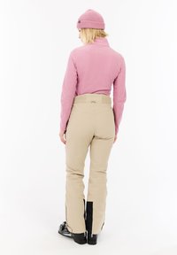 Felpa in pile a maniche lunghe di color rosa chiaro, abbinata a pantaloni da neve beige con vita elastica e cerniere laterali nere. Stivali da sci neri visibili.
