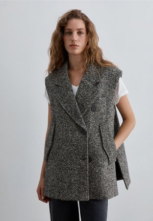 Mujer con cabello ondulado lleva un chaleco de tweed gris sin mangas sobre una camisa blanca de manga corta, parada frente a un fondo claro y liso.