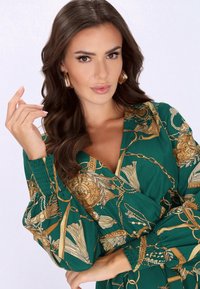 Vestido envolvente verde con cadena dorada y patrón floral, mangas globo con puños fruncidos y escote profundo en V. Pendientes de aro dorados.