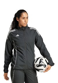 Schwarze Adidas Sportjacke mit weißen Streifen, Reißverschlusschluss und hohem Kragen. Das Modell hält einen schwarz-weißen Fußball und trägt passende Hosen.