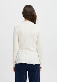 Camiseta de manga larga de punto en color crema con un escote y dobladillo con forma de ondas, que presenta un diseño ajustado, una textura suave y un patrón de rayas verticales.
