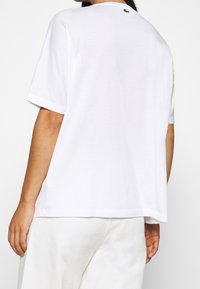Camiseta de algodón blanca con mangas cortas, corte holgado y un pequeño detalle de logo verde en la parte posterior del cuello. Textura suave, diseño minimalista.