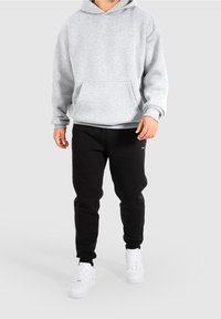 Sweat à capuche gris avec poche avant, porté avec un pantalon de survêtement noir et des baskets blanches. Tissu doux, coupe ample et détails de couture visibles.