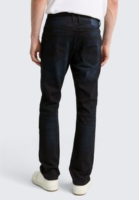 Homme portant un jean bleu foncé et des baskets blanches, se tenant de dos face à la caméra sur un fond clair et uni.