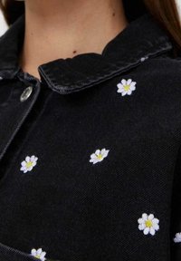 Czarny denimu ubranie z kołnierzykiem, ozdobione białymi haftowanymi stokrotkami z żółtymi środkami, rozsianymi po tkaninie.