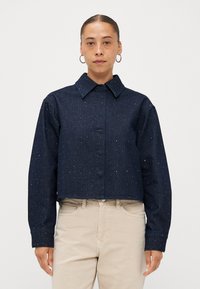 Korte donkerblauwe spijkerjack met een kraag, versierd met verspreide lichte embellissementen. Lange mouwen met knoopsluiting bij de manchetten.