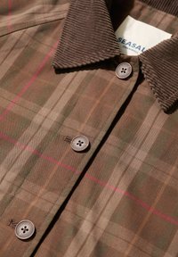 Brūns šķērsgriezuma krekls ar korduroja apkakli, diviem brūniem pogām, horizontālām un vertikālām līnijām zaļā un rozā krāsā. Marķējums redzams iekšpusē.