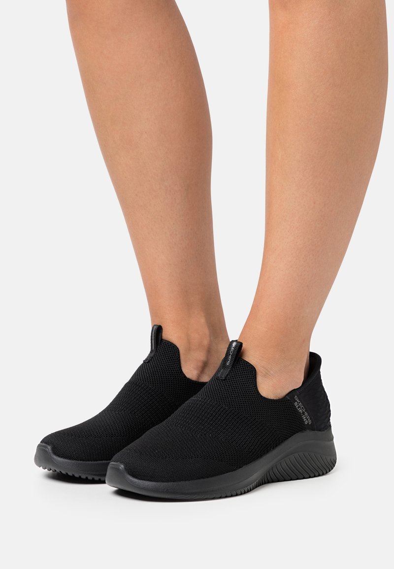 Deux jambes portant des baskets en tricot noir sans lacets avec des semelles texturées sur un fond clair uni.
