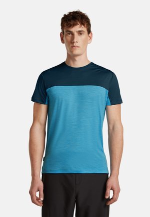 Mann, der nach vorne schaut, trägt ein kurzärmeliges, zweifarbiges blaues T-Shirt und schwarze Hose vor einfachem Hintergrund.