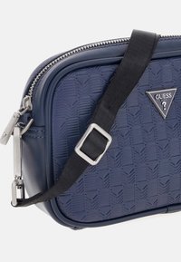 Bolso cruzado de color azul marino con patrón geométrico en relieve, herrajes plateados y correa negra. Cuenta con cierre de cremallera y detalle del logo de la marca.