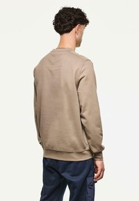 Mann mit lockigem, dunklem Haar, der einen beigen Sweatshirt und marineblaue Cargo-Hosen trägt, mit dem Rücken zu einer einfarbigen, hellen Hintergrund.