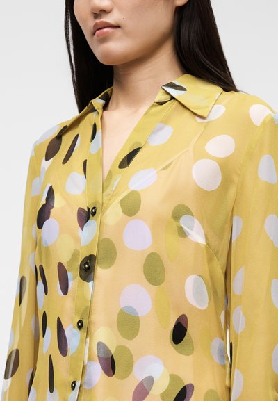 Πουκάμισο με μακριά μανίκια και κουμπιά σε κίτρινο ύφασμα, με διαφανή υφή και πολύχρωμες πουά σε διάφορα μεγέθη και αποχρώσεις.
