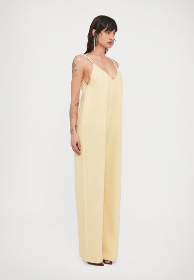 Γυναίκα που στέκεται πλάγια, φορώντας φαρδιά ολόσωμη φόρμα σε απαλό κίτρινο χρώμα με λεπτές τιράντες και σκουλαρίκια, μπροστά σε λευκό, απλό φόντο.
