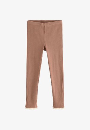 Smilende pige står iført en hvid sweatshirt med blomsterprint, brune ribstrikkede leggings og mørkebrune flettede sko mod en ensfarvet baggrund.