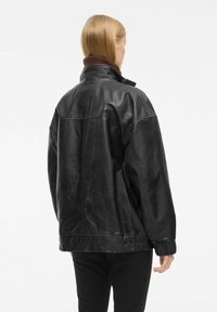 Veste en cuir noir avec un col montant, présentant une finition texturée et des poignets élastiques. Le design est ample avec des coutures visibles.