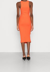 Obepínající žebrované šaty v jasně oranžové barvě, s bezramínkovým střihem a délkou po kolena, kombinované s černými sandály s otevřenou špičkou.