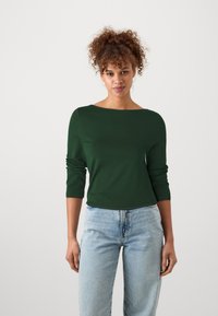 Dark green long-sleeve top with a boat neckline, made from smooth fabric; paired with light blue, loose-fitting jeans.