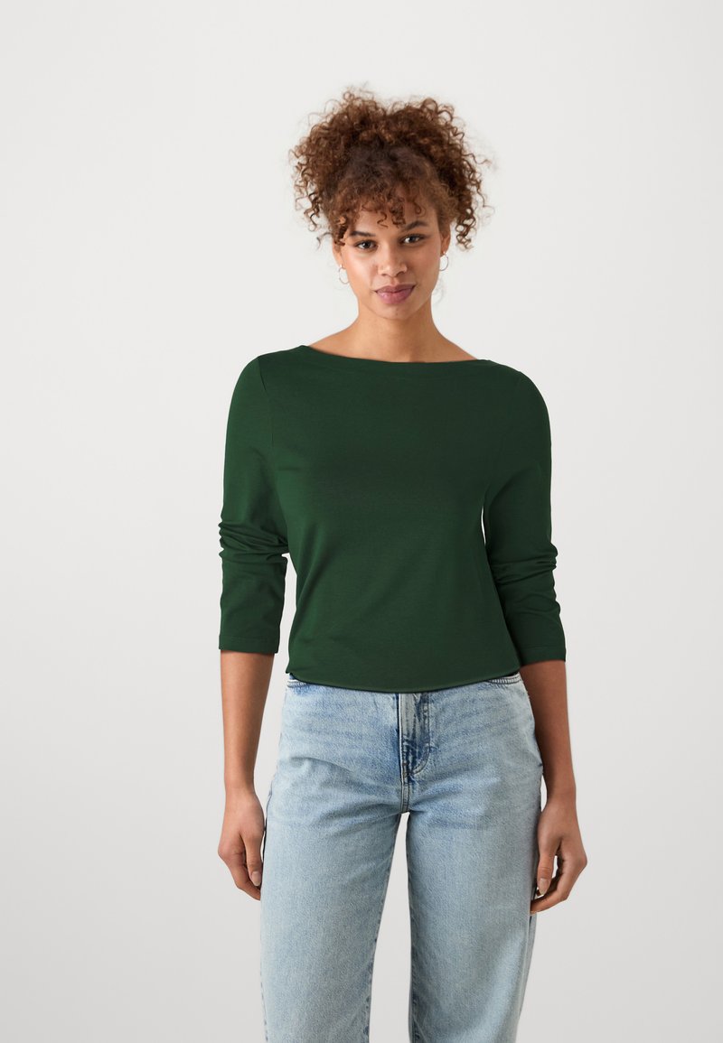 Dark green long-sleeve top with a boat neckline, made from smooth fabric; paired with light blue, loose-fitting jeans.